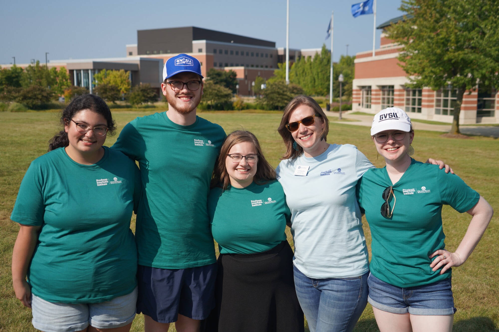 A group of five people posing for a photo - four of them are students wearing matching teal shirts with their staff advisor on the GVSU lawn.